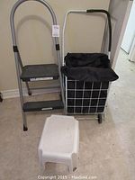 Photo showing the fold up step ladder with metal frame, two black steps and curved handle, fold up cart with white wire frame and black fabric basket, and small white plastic step stool nearby.