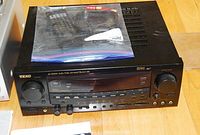 Front and top view of the black TEAC AG-D9100 audio/video surround receiver on a wooden floor with a Ziploc bag containing the remote control and other accessories on top.