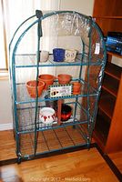 Mini greenhouse with 4 shelves and various ceramic and terracotta plant pots inside, clear plastic cover and green metal frame on wheels