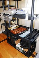 Three-tier black plastic shelving unit with various kitchen items including warming tray, knife set, roasting pan and mason jars on shelves