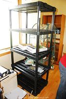 Full view of tall black heavy plastic shelving unit with five slatted shelves, placed near window.