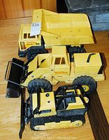 Three vintage Tonka construction vehicles shown together on a wooden floor: dump truck, front end loader, bulldozer.