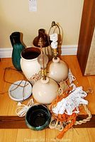 Pair of spherical cream ceramic lamps with cords and various pottery pieces grouped on a hardwood floor including vases, jug, small bowl, and macramé hangers.