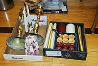Photo showing two large brass candle holders, two silver-colored handled candle holders, and a box with assorted candles including tapers and tealight candles in multiple colors.