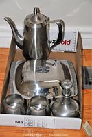 Stainless steel covered dish with Oneida coffee pot, cream, sugar, and pewter cruet on a wooden surface with a white background.