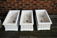 Front view of three white rectangular concrete planters with fluted designs and drainage holes, arranged outdoors against a brick wall.