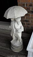 Full frontal view of concrete fountain showing boy and girl standing under an umbrella, girl holding a teddy bear.