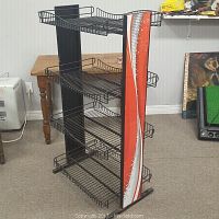 Front angled view of black metal wire display rack with four basket shelves and Coca-Cola branded side panels showing swirl and bubble graphic in red and white.