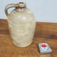 Stoneware crock jug with handle and narrow opening, shown next to a measuring tape for scale, approximately 10 inches tall, no visible chips or cracks.