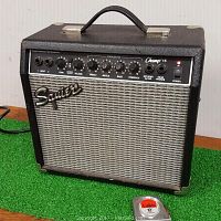 Front and left angled view of amplifier showing Squier branding, control knobs, and handle on top, placed on green carpet with measuring tape next to it for scale.