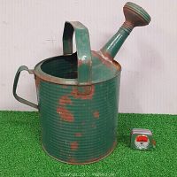 Side view of the green vintage tin watering can with rust spots, showing large arched handle, smaller back handle, and attached spout with watering head; measuring tape for scale.