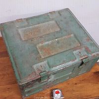 Top angled view of a square green metal box with rust, stamped with 'C 1944 OMS/C' on the lid. A tape measure is placed nearby for scale.