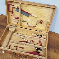 Wooden toolbox open to show vintage kids metal tools arranged inside; includes saw, clamp, wooden hammer, level, hand drill and broken pliers.