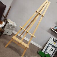 Front view of the wooden easel standing on carpeted floor in a room, showing height and frame structure with adjustable supports.