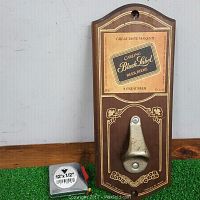 Front view of wooden plaque featuring Carling Black Label print with metal bottle opener mounted below, measuring tape visible for scale.