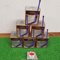 Six unopened Crown Royal whiskey glasses in limited edition packaging stacked on green turf with a purple hockey stick shaped stir stick next to them, alongside a tape measure for scale.
