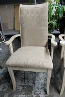 Front view of a newly upholstered beige dining armchair with armrests and tall back, showing swirl pattern on fabric and white wooden frame.