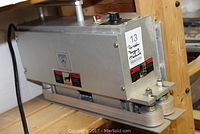 Side view of Emplex portable bagging machine showing the metallic body, rollers, and control knob.