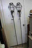 Full view of two tall metal garden torches leaning against a door, featuring geometric cage tops and long stakes, with some finish wear visible.