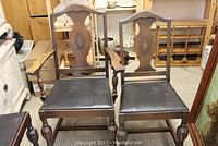 Photo showing 4 wooden chairs with black upholstered leather seats. One chair is an armchair with armrests. Chairs have carved wooden backs with central decorative motifs. Visible wear consistent with age.