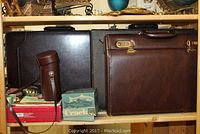Three briefcases: dark brown leather with lock, black, and new-ish brown briefcase. Fishing rod case and Crack fishing reel box in front.