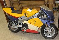Side view of motorized pocket bike showing yellow and blue plastic fairing, red lower fairing, black foot pedal, front wheel and brake disc.
