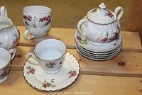 Photo showing stacked teapot, several saucers and cups, and a creamer, all with floral rose pattern and gold trim.