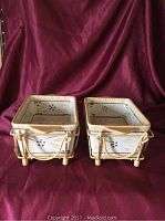 Two ceramic loaf bakeware pans in wire holders side by side, showing decorative patterns and lids on top.