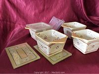 Four square ceramic ovenware dishes with plastic lids and two glass table protectors on display on reddish cloth background.