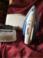 White and blue Panasonic steam iron standing vertically with adjustable settings visible and white plastic carrying case in background.