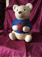 Front view of a ceramic teddy bear cookie jar featuring a cream color, blue sweater, and red heart held by the bear.