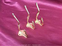 Three wall-mounted brass coat hooks displayed in a row on a red fabric background, showing the front with two hooks on each and the mounting holes.