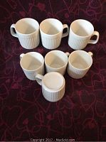 Seven ceramic coffee mugs arranged upright and one mug placed upside down showing the base. White mugs with ribbed Athena pattern.