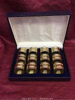 Opened box showing 12 brass napkin rings neatly arranged in 3 rows on blue velvet insert.