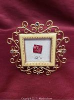 Front view showing brass-colored frame with ornate design, enamel mat, and rhinestones around a 2x2 inch placeholder.