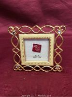 Front view of the brass-colored square photo frame with enamel mat and rhinestone accents against a red fabric background.