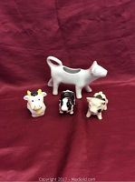 Four ceramic cows displayed on a red fabric background, including a milk pitcher cow and three smaller cow figurines in different colors and patterns.