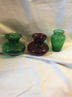 Three small glass vases, two green and one red, displayed on a white fabric background.