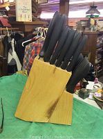 Knife block containing 18 knives with black handles in a wooden holder, placed on a green cloth.