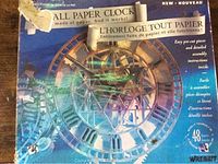 Box front showing the assembled paper clock model against a blue sky background. Text highlights the model is entirely made of paper and functional.