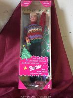 Front view of Barbie doll in original packaging showing festive sweater, box graphics, and included accessories.