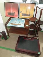 Photo showing three framed photographic prints of ships mounted on a wall or leaning, with a wooden plant stand, a Georgian mahogany end table, Edwardian mahogany writing table below, and a dark wooden matchstick box on the floor.