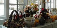 Wide view of assorted artificial flowers in vases, metal watering can, ceramic urn, wooden cow bookends, stained glass ornaments and fruit display items on a table.
