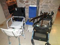 Overview of lot showing white shower chair with armrests, blue toilet assist frame, blue and black walkers with seats and baskets, wheelchair partially visible in background, and several canes.