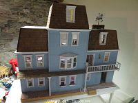 Exterior front view of the light blue painted wooden dollhouse with brown shingled roof and white balcony