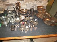 Full overview of assorted silver plate and pewter serving pieces on table showing trays, teapots, candlestick holders, napkin holders, salt cellar, and cups.
