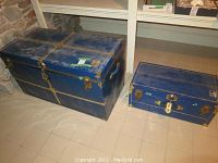 Two blue metal shell trunks side by side, showing exterior and hardware with patina and wear.