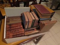 Top-down view of a collection of 14 old books packed in a shoebox on a table; various bindings visible including leather and cloth, some with noticeable wear and aging.