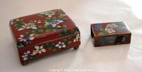 Photo of cigarette box and match holder side by side showing floral cloisonne enamel decoration on reddish background.