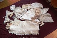 Large assortment of antique crochet and needlework linens spread out on a dark fabric background, showing various sizes and patterns, including trimmed linens and doilies, as well as a large rectangular tablecloth.
